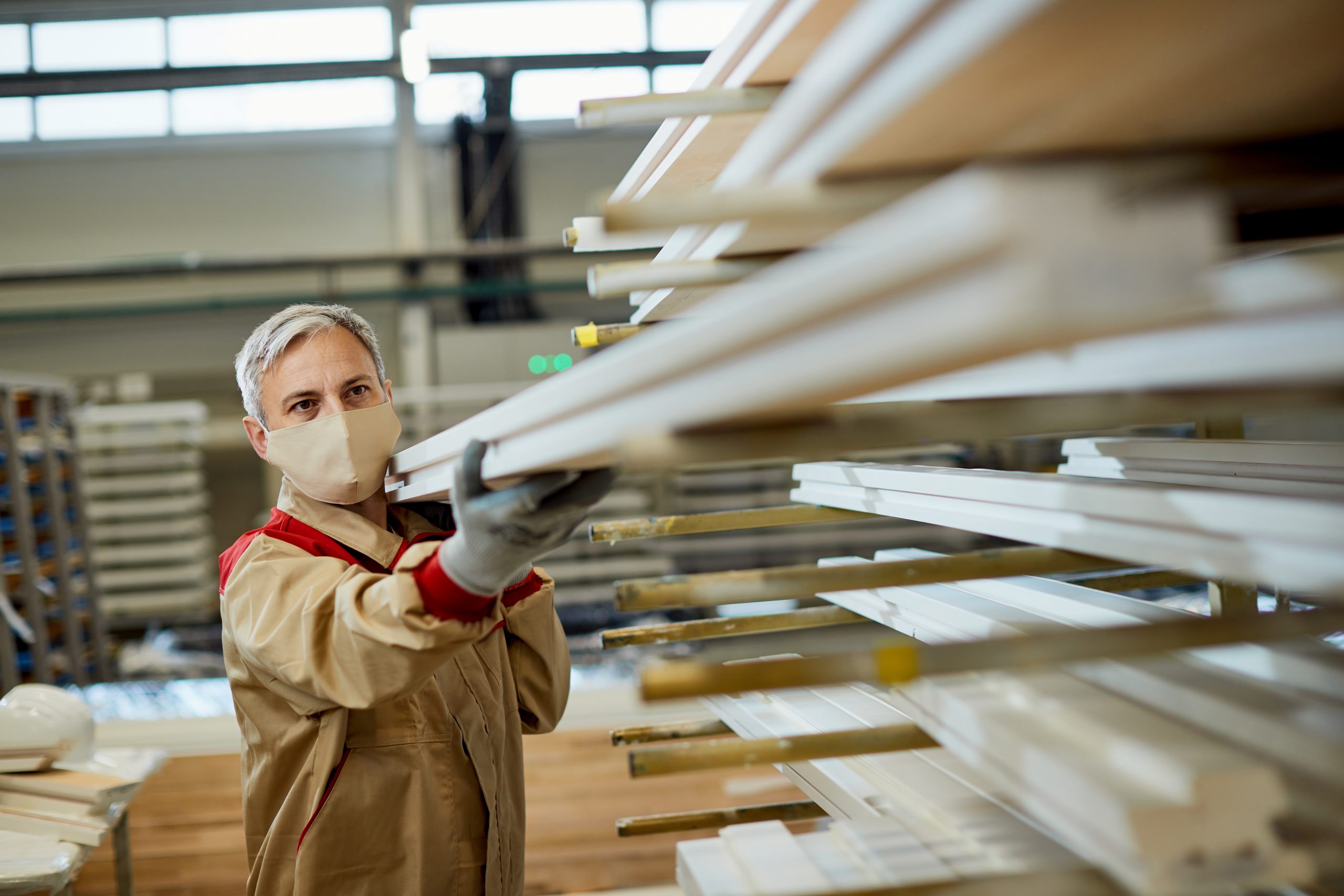 Male carpenter wearing protective face mask while taking wood planks from a shelf in a workshop.