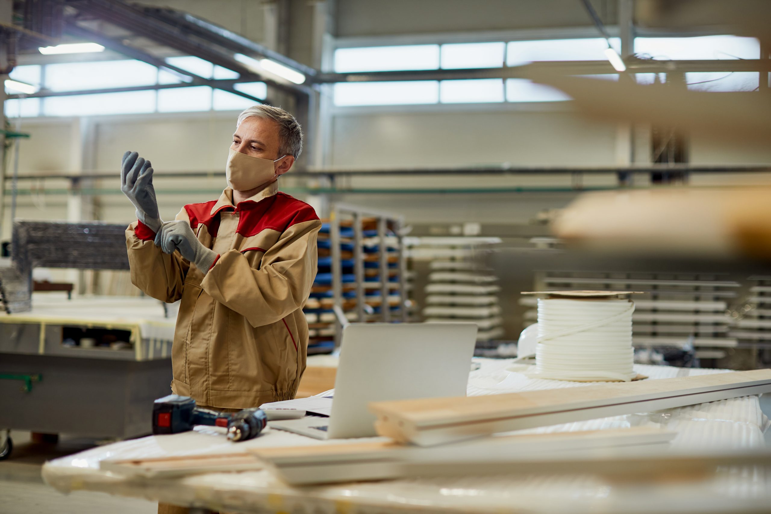 Mid adult carpenter wearing face mask and putting on working gloves in a workshop.
