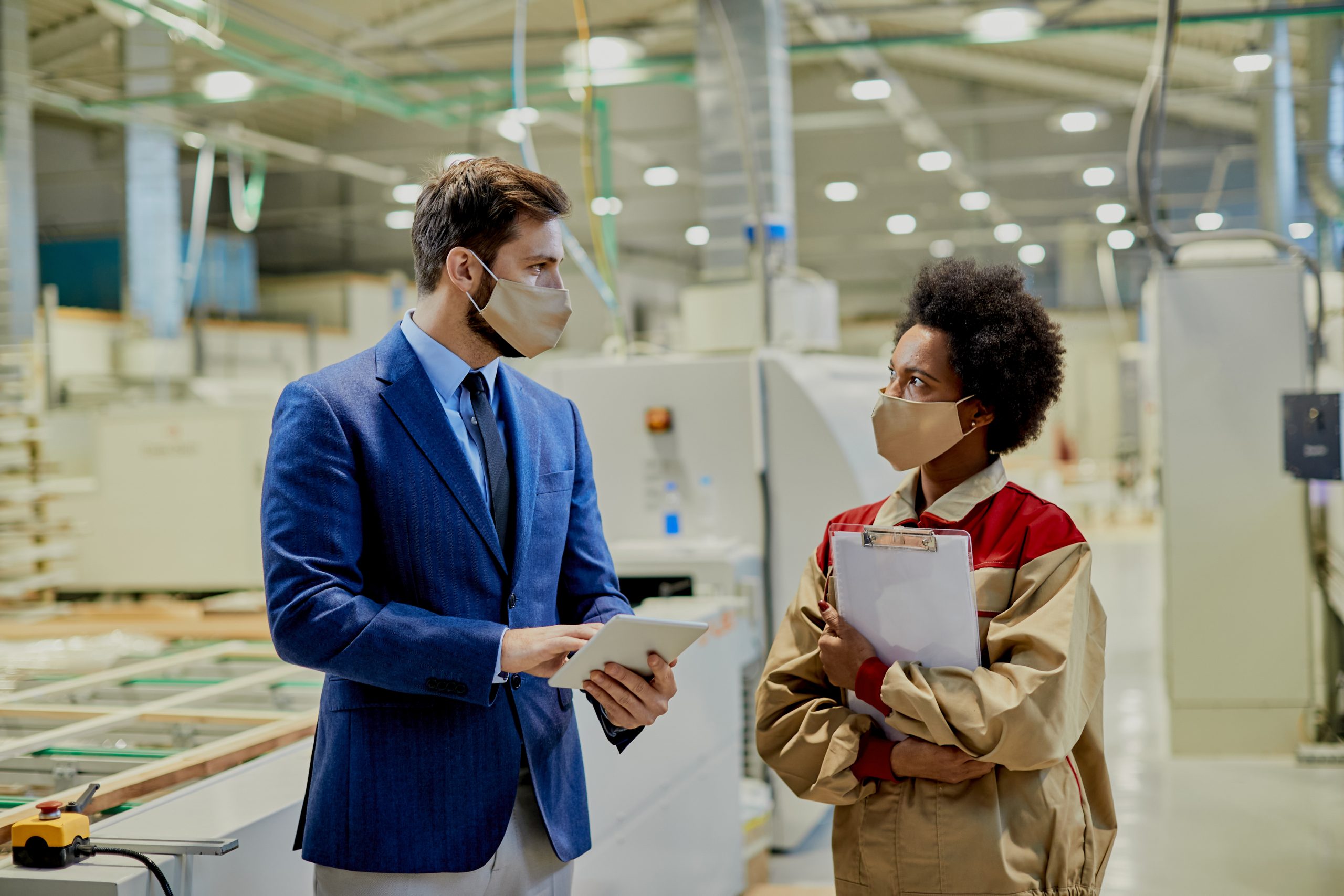African American factory worker and company manager wearing face masks while communicating at woodworking production facility.